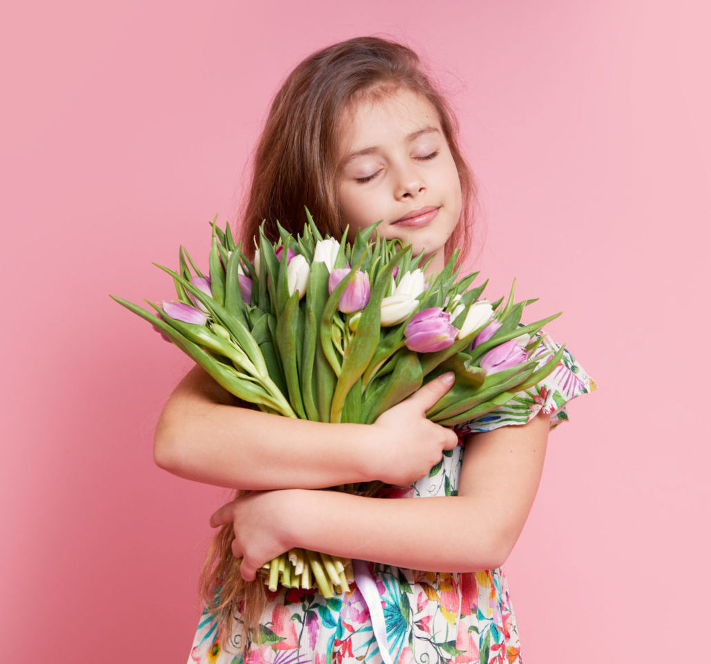 Cute smiling child girl holding bouquet of spring flowers tulips isolated on pink background. Little toddler girl gives a bouquet to mom. Copy space for text.